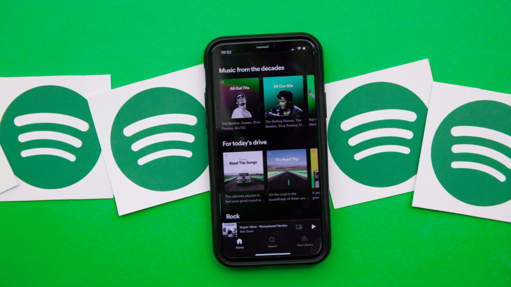 Spotify shade green background. Mobile phone with Spotify app up in the middle of the page. Two Spotify logos on white square backgrounds either side of the mobile.