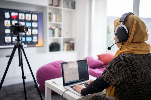 Woman with headphones on, looking at laptop screen with a camera on a tripod set up in front of her and a TV screen ahead with lots of apps. She is sat in a room with book shelves on the right of the TV screen and pink beanbags.