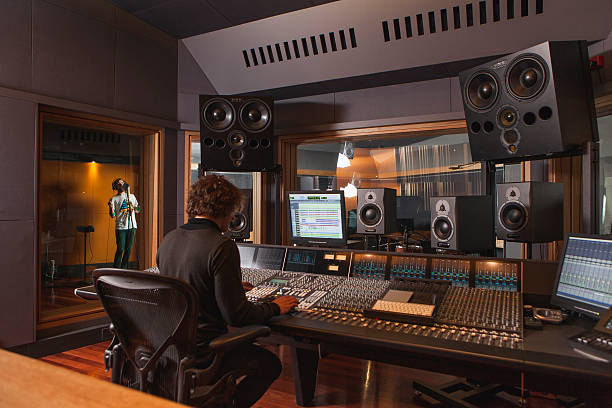 Studio set up, a music producer sat in front of equipment with a artist singing in a room to the left of him.