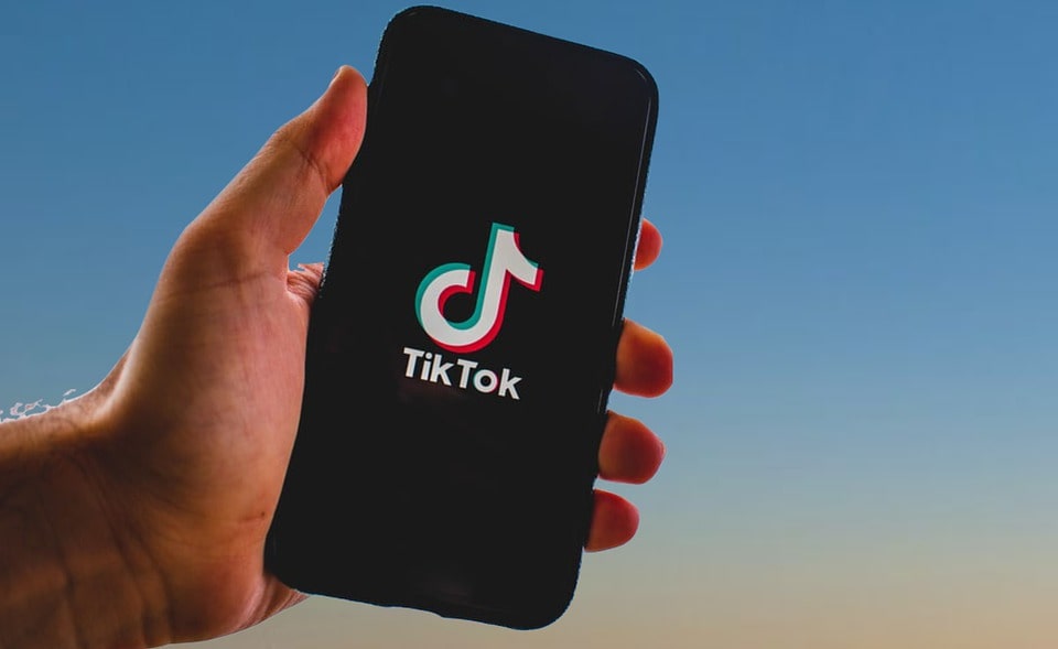 Blue to pink gradient effect background. Foreground has a male hand holding a smartphone with the TikTok logo on it.