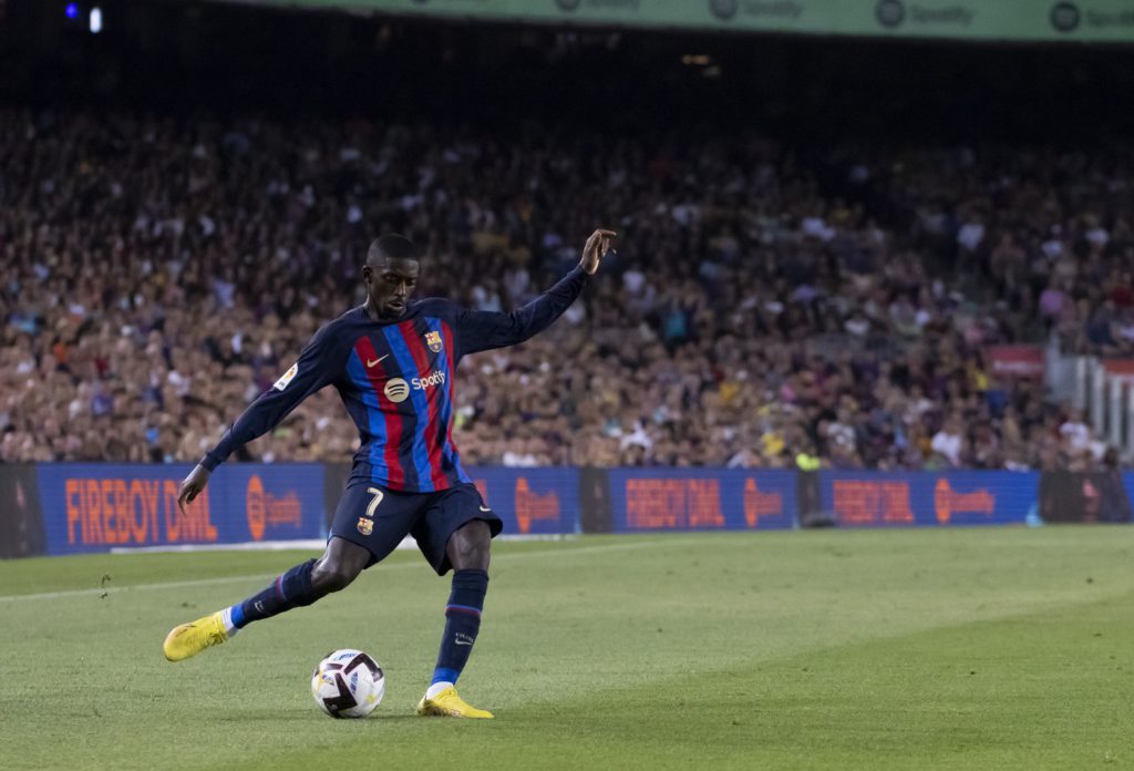Ousmane Dembélé preparing to kick a football. In the background by the crowd is an LED sign advertising Spotify and FIREBOY DML