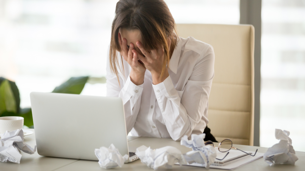 Girl with head in her hands sat at a desk in front of her laptop with paper scrunched around her.