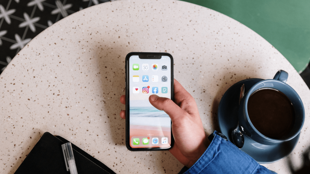 Image of a table with a cup of black coffee and a pen. Someone's hand is holding a smartphone over the table, displaying their apps. One of which is Instagram.