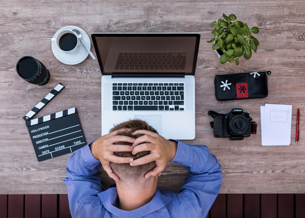 Image of man with his hands on his head while sat at his desk appearing stressed. Surrounding him is his camera, a clapperboard and his laptop.