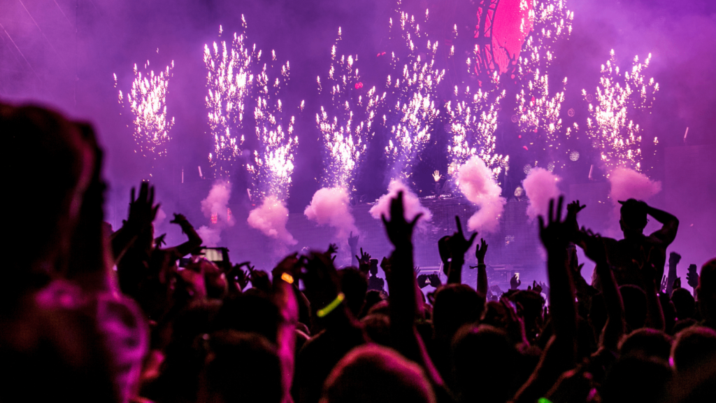 Purple smoke and fireworks on stage. In the foreground is a crowd of people.