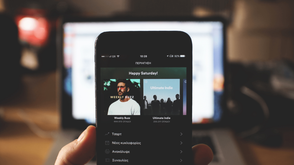 Smartphone with Spotify loaded. Behind the phone is a laptop on a desk.