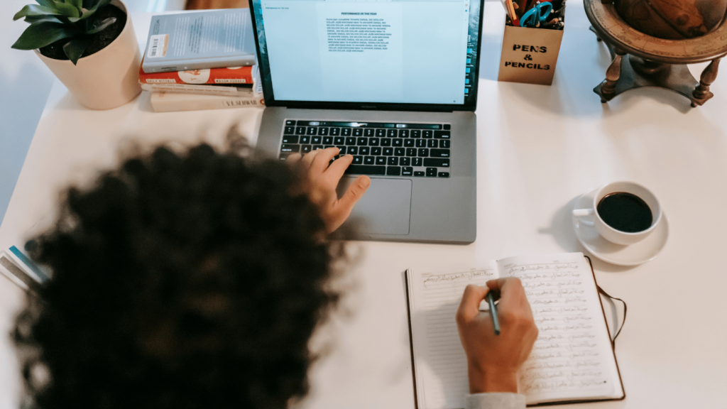 Woman sat at desk with a pen in her right-hand writing and her left hand resting on her laptop keyboard. On the desk are a coffee, plant, books, globe and a pen pot.