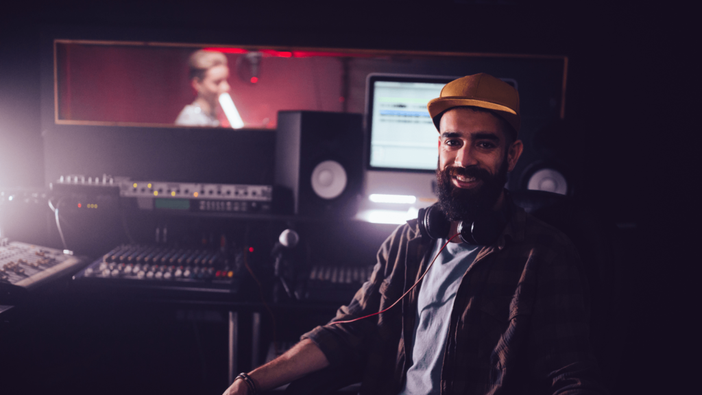 Man smiling at the camera sat in front of recording equipment. Someone in the background is singing into a microphone.