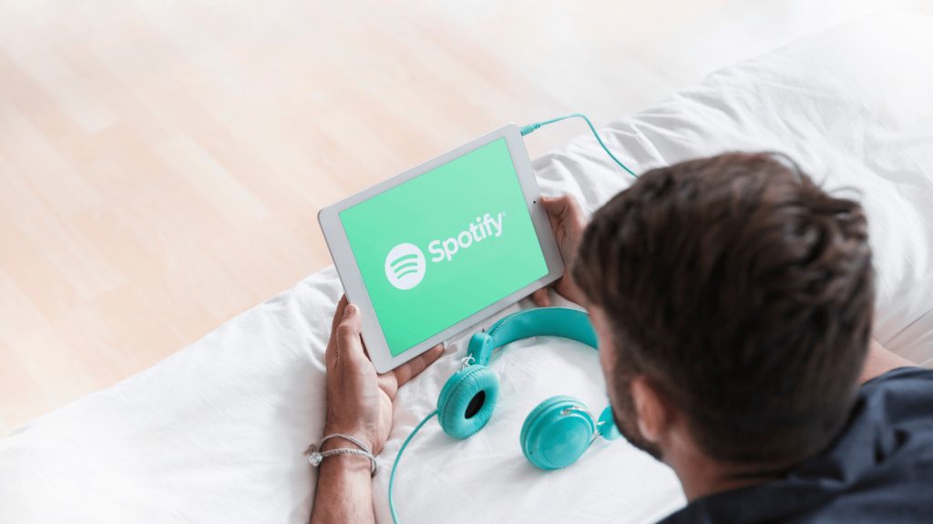 Man lay on a bed with headphones next to him, viewing the Spotify logo on a tablet.
