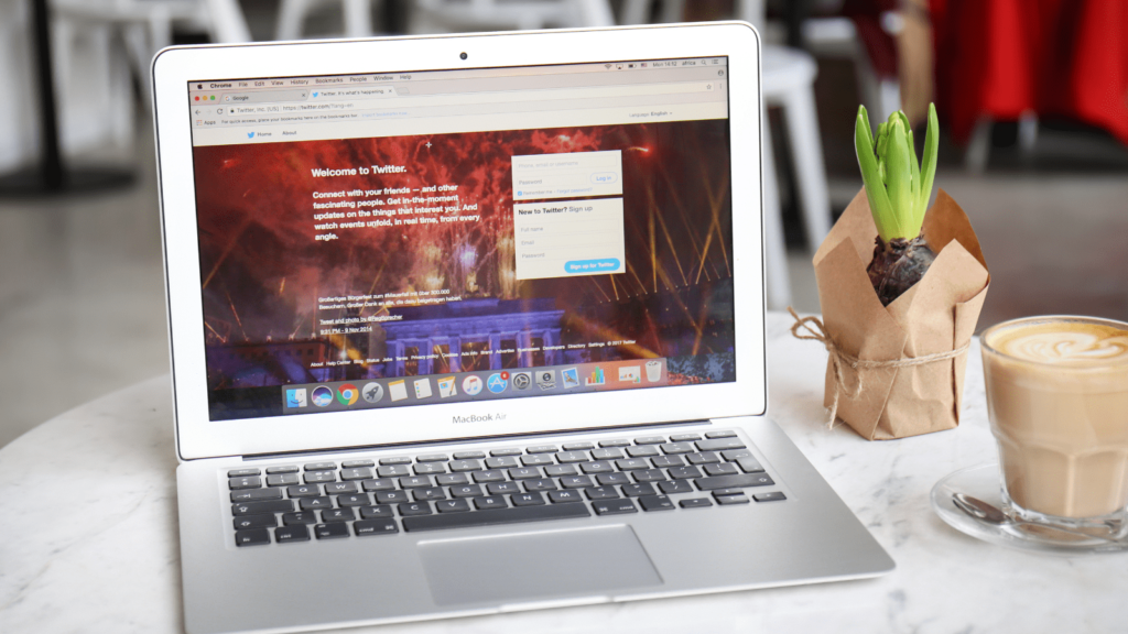 Twitter loaded on a laptop. Laptop is placed on a desk with a plant and a coffee.