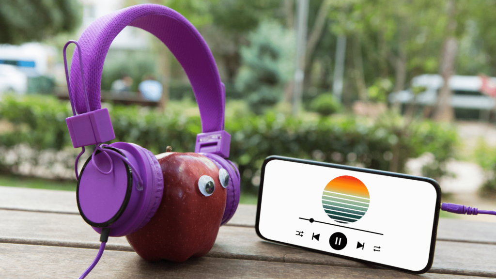 An apple with googly eyes wearing headphones plugged into a smartphone that is displaying an album.