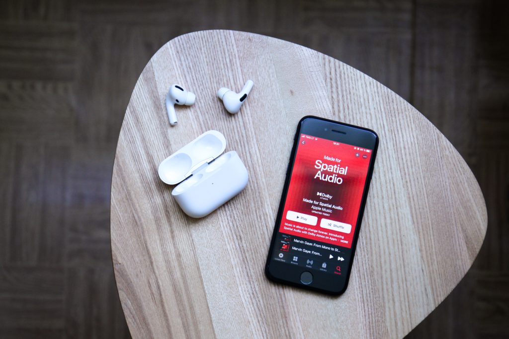 Apple Music to pay higher royalties for artists creating spatial audio. Photo of a smartphone next to Apple earbuds. On the phone, a Spatial Audio playlist is loaded.