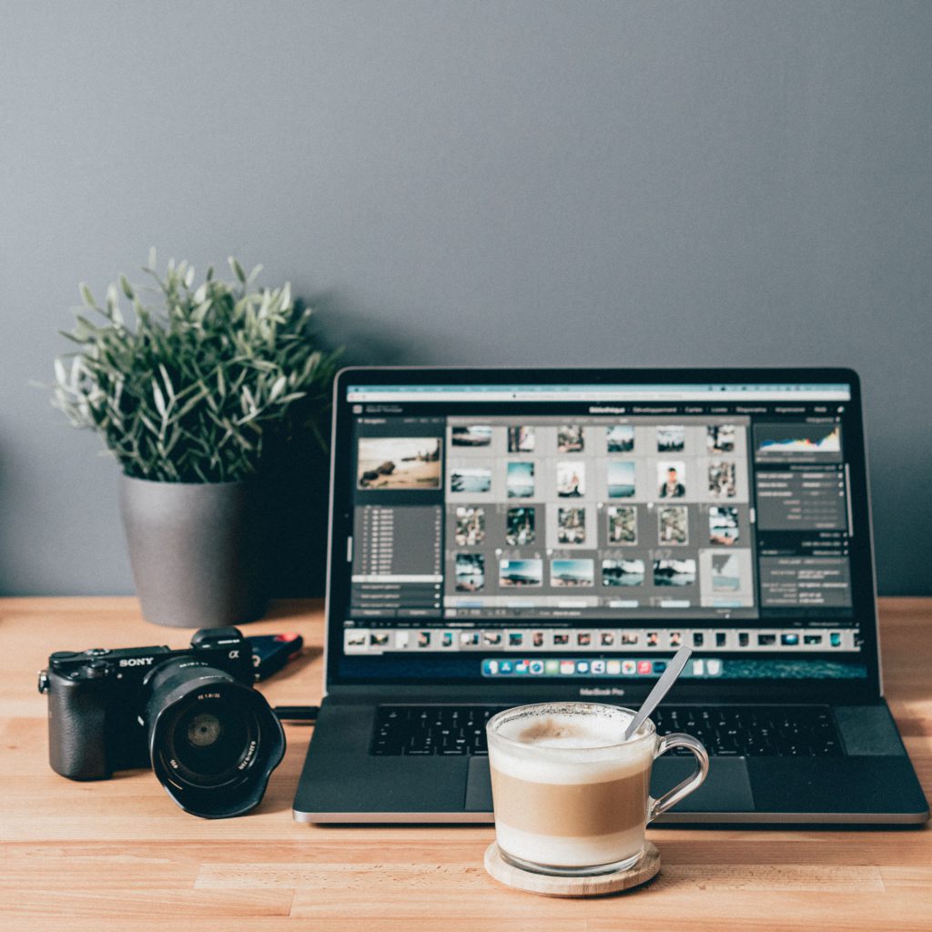 How to easily choose photo editing software. Photo of a grey wall and wooden worktop. On the worktop is a plant in the background. In the foreground is a laptop with photo editing software loaded, a camera and a coffee.