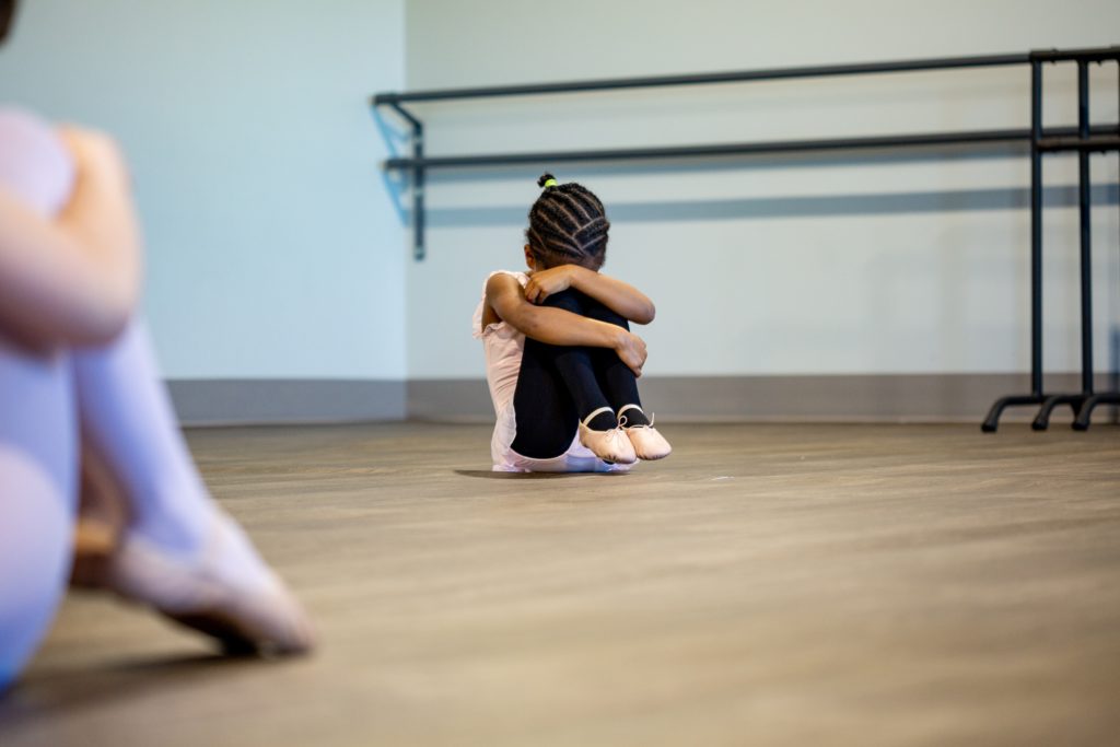 Conquering stage fright - embrace the spotlight with confidence. Photo of a little girl curled up with her knees to her head in a ballet outfit, sat on the studio floor.