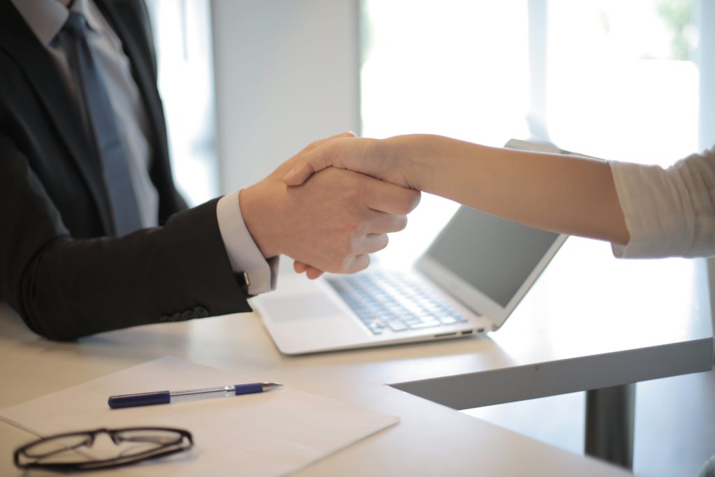 First impressions count - how to land your perfect job. Photo of two people shaking hands. You can't see their faces, but can see a desk with a laptop, pen, paper and glasses on.