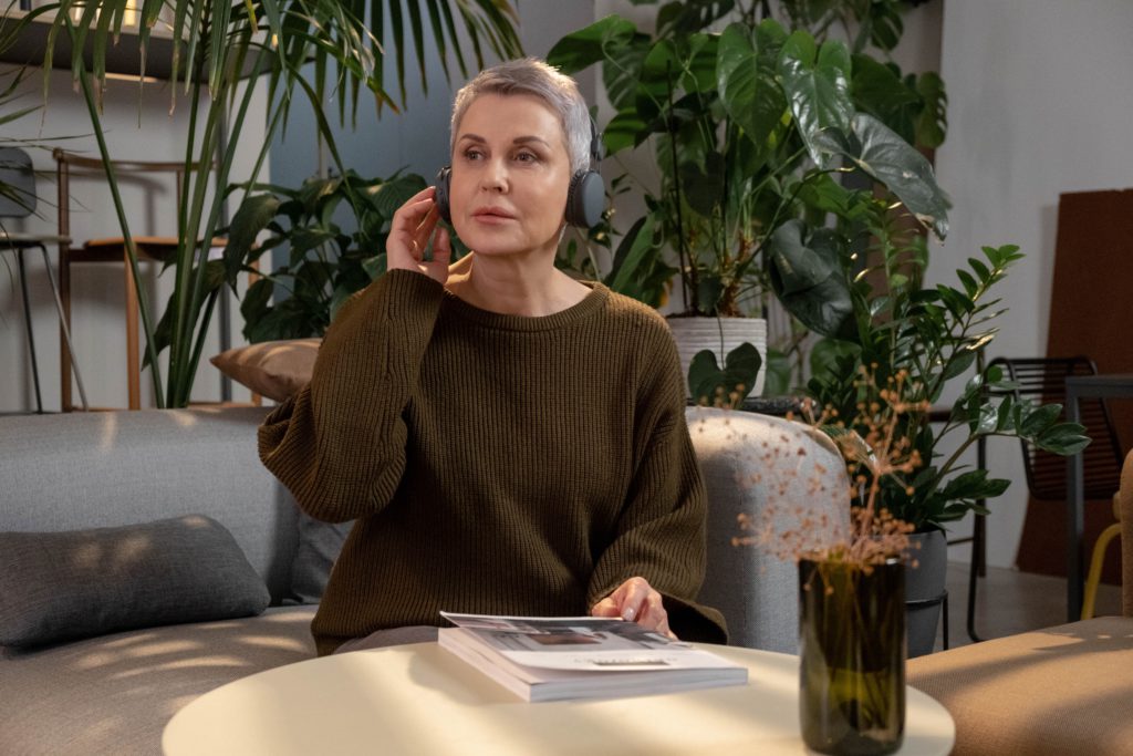 What is Spatial Audio and why is it so popular? Photo of a woman holding one side of her headphones. She is sat on a sofa in a clean, modern looking room surrounded by plants.