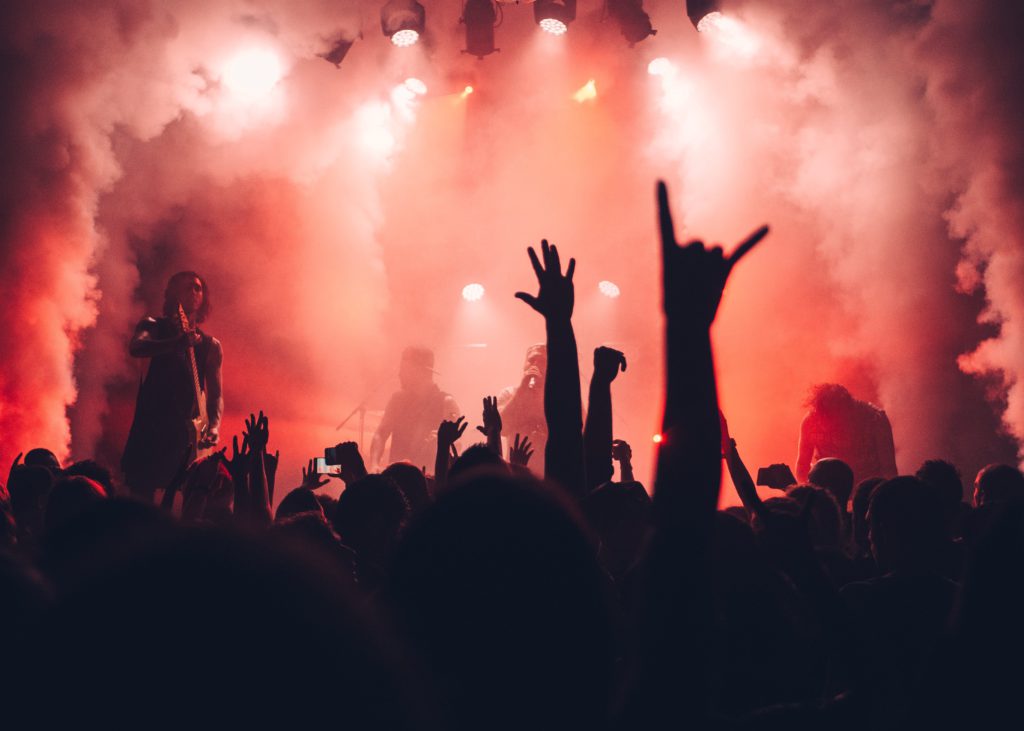 3 websites for music artists to find free classic rock samples. Red smoke filled stage with musicians on it. Close up of crowds silhouette, one person is throwing a rock on sign with their hands.