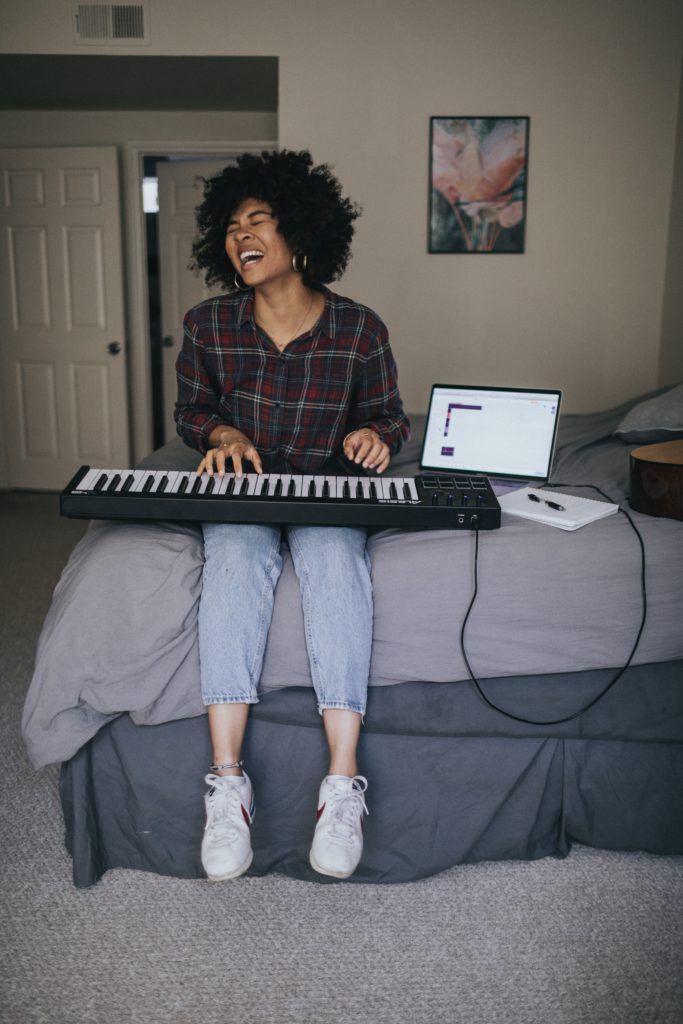 5 side hustle ideas for independent musicians. Photo of a girl sat on her bed singing, while playing the keyboard plugged into her laptop.