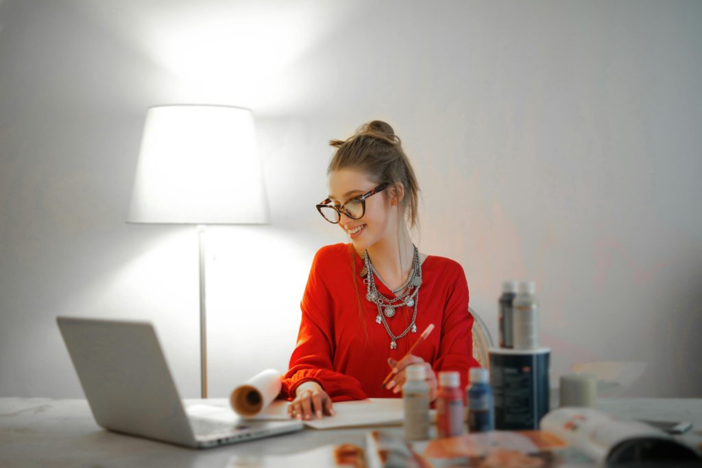 Freelancing mysteries - what exactly is a freelancer? Photo of a woman looking at her laptop screen, smiling, with a lamp behind her and paints in front of her.
