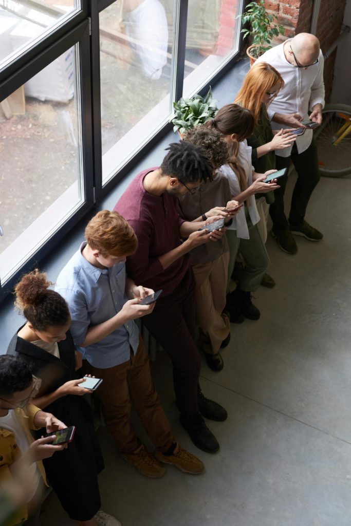 The origin story - where social media first started. A line of people stood side by side against a window. They're all looking at their smartphones.