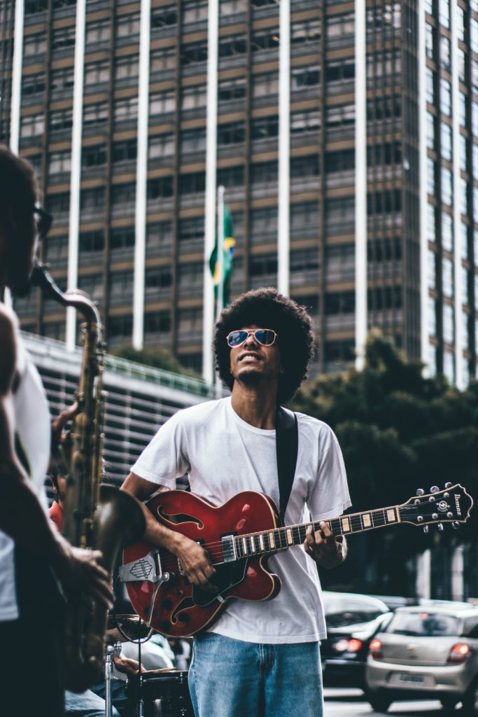 Debunking common myths about music artists. Photo of a man with an afro playing his guitar in a city centre, next to someone with a saxophone. 