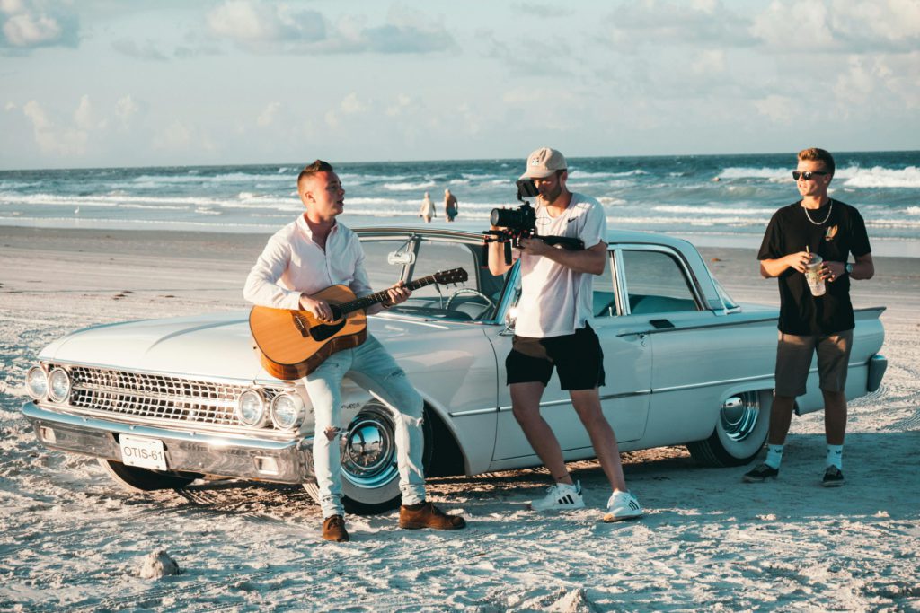 The power of visual - do small music artists need music videos? Photo of three men on the beach, one is sat on a car with his guitar and another is recording it.