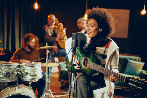 4 reasons a musician should never quit their passion. Photo of a girl singing into a microphone, holding a guitar. In the background is a man on drums, another on a trumpet and another you can't see too well.