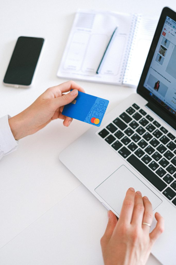 Bringing your online business into the physical world - a step-by-step guide. Photo of a hand holding a credit card while hovering over a laptop. Next to the laptop is a smartphone, paper and a pen.