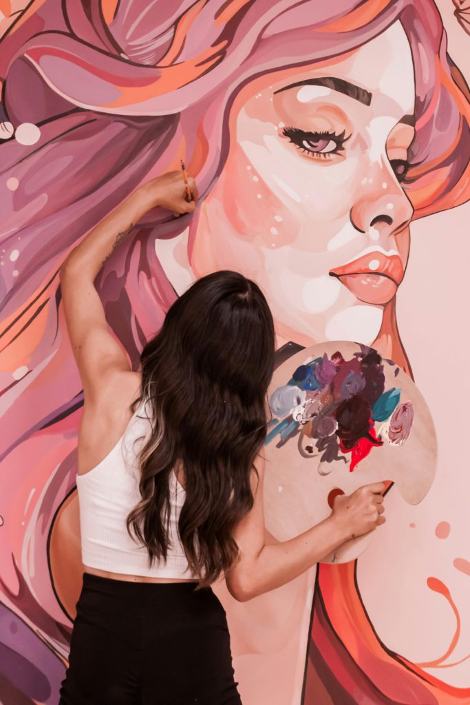Embracing sustainability - the rise of eco-friendly art and design. Photo of the back of a girl. She is painting a huge woman's face on a wall. She is holding her paint palette in her right hand and her paintbrush in her left hand.