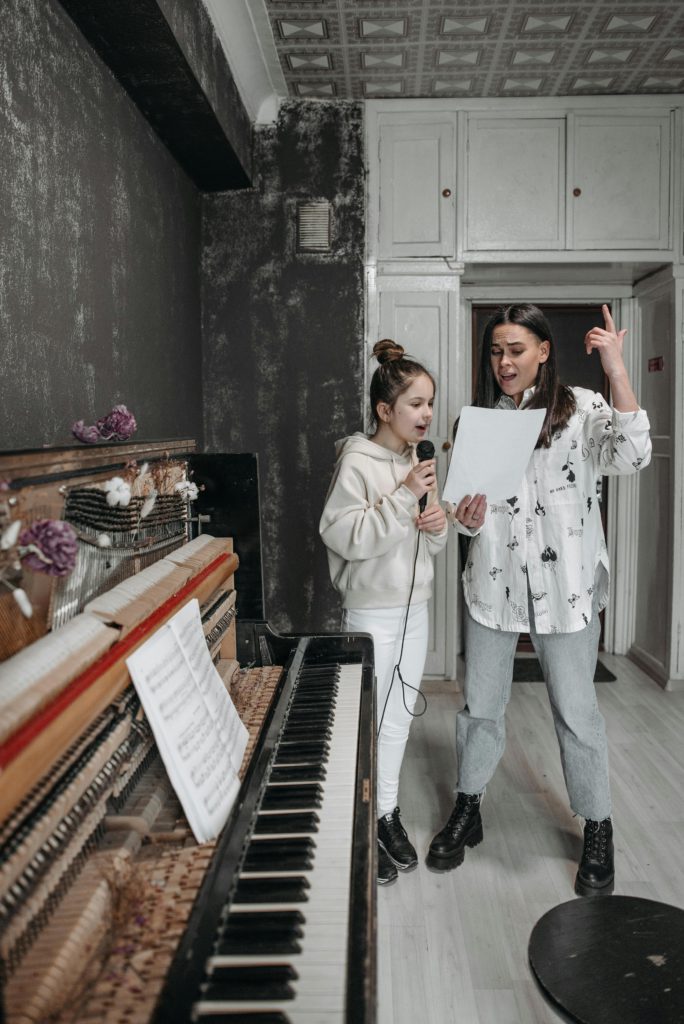 How to add lyrics to your music on Spotify. Photo of a woman and a girl in a room near a piano singing into a microphone while reading from a song sheet.