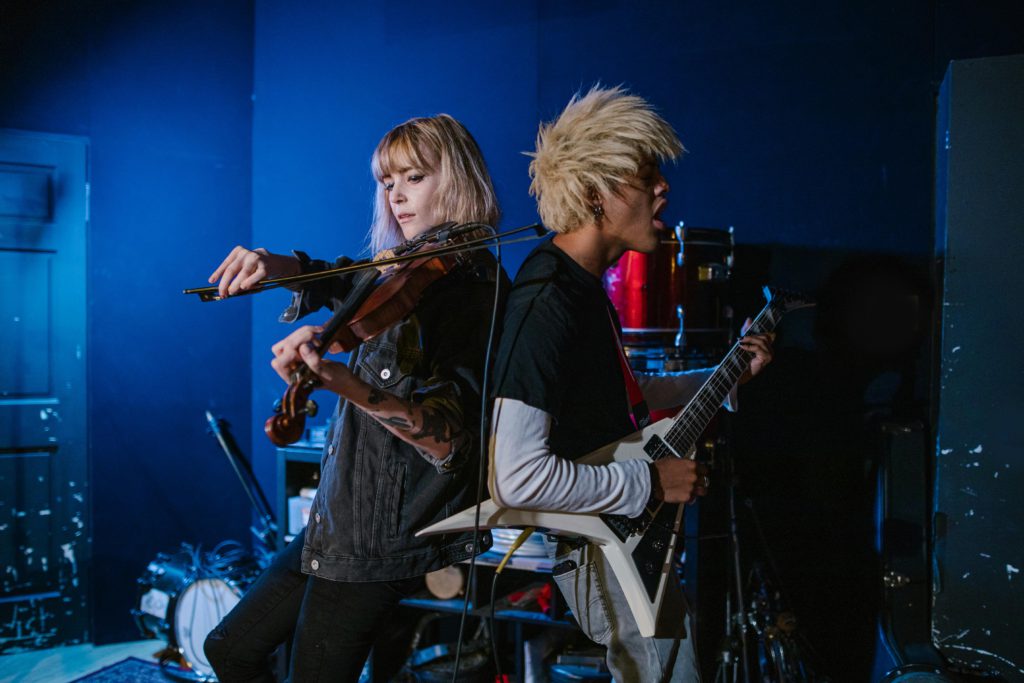 Behind the scenes - how music distribution platforms work. Photo of a female playing the violin with her back to a male playing the electric guitar while singing.