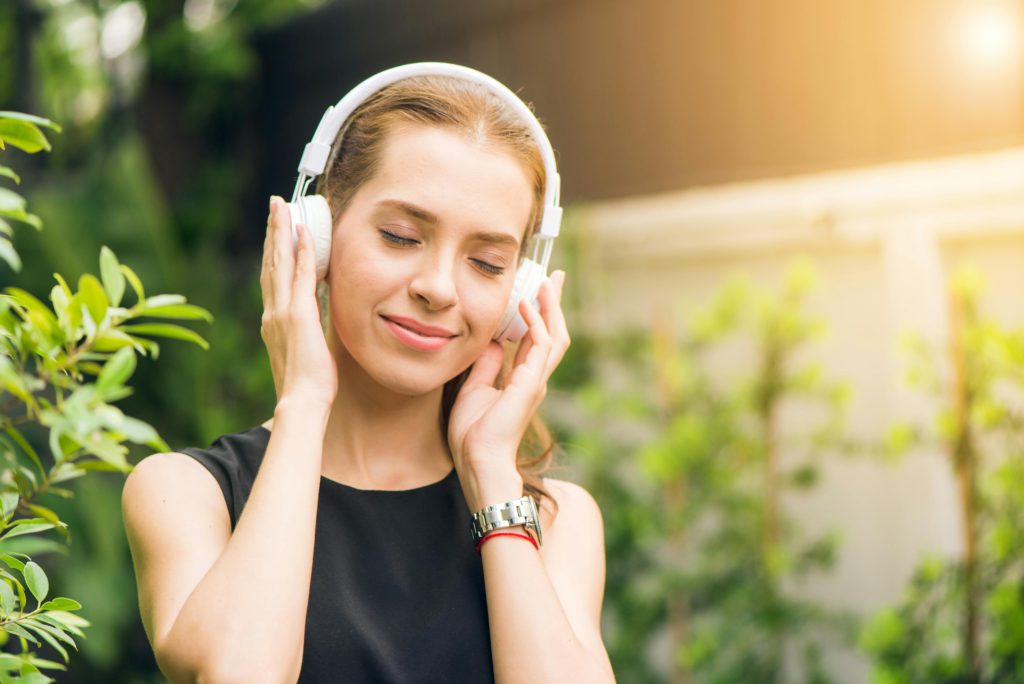 Exploring the healing power of music therapy - how music can improve mental well-being. Photo of girl holding headphones to hear ears, smiling outside.