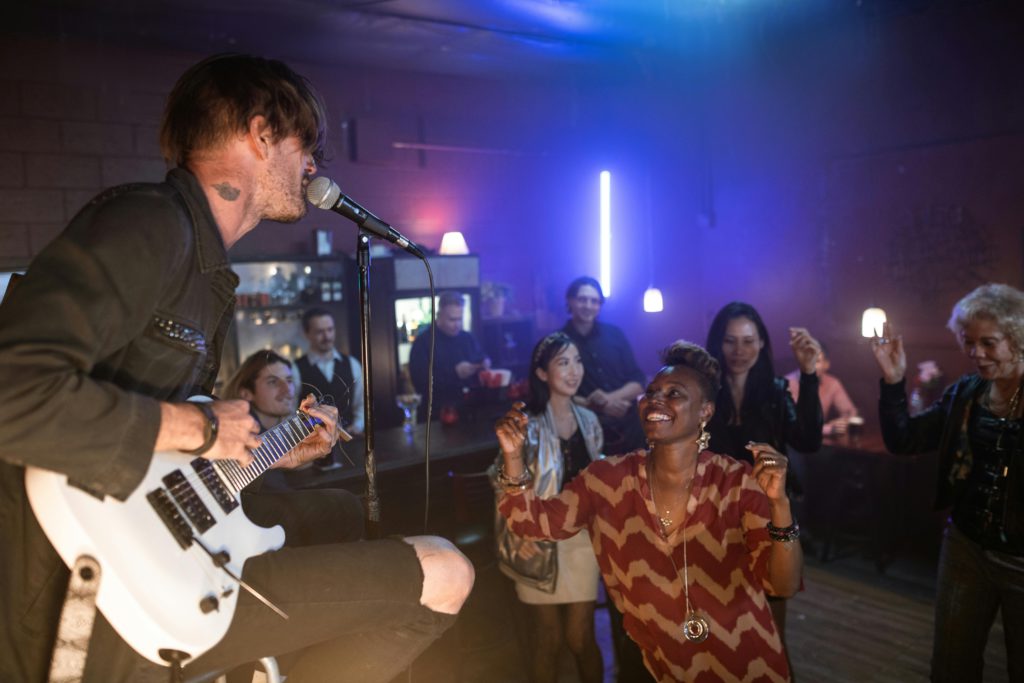 Easy performance tips for musicians. Photo of a male singing into a microphone while playing the electric guitar. A woman has stepped forward from the dancing crowd, smiling.