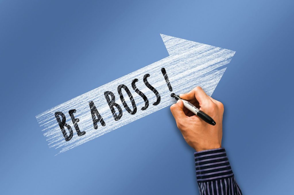 Becoming your own boss - what you need to learn. Photo of a blue background with a white arrow. Someone's hand writing "Be A Boss!"