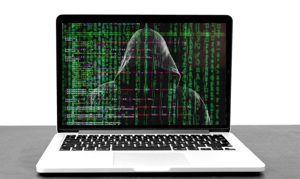 What to do if you believe your Facebook has been hacked. Photo of a laptop and code is loaded onto it. In the background, you can see a person in a hooded cloak.