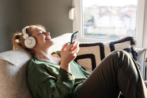 Spotify Instagram notes integration in the works to share real-time listening activity. Girl smiling with her eyes closed as she sits on the sofa listening to music through her headphones with her phone in her hand.