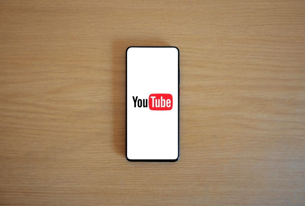 YouTube updates - collab playlists, fan badges and sleep timer. YouTube logo on a smartphone against a wooden background.